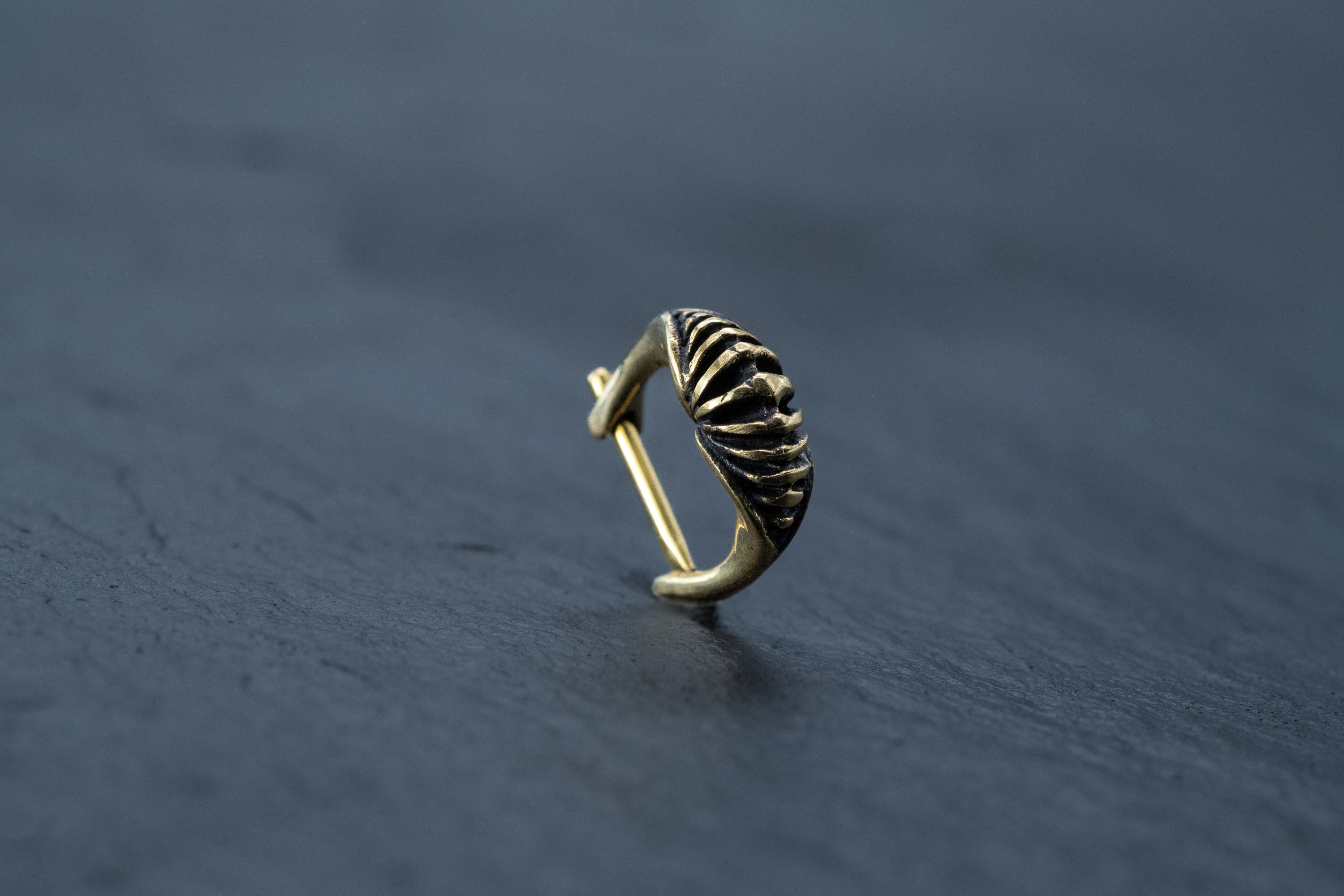 A close-up image of the DIARA Biomechanical Clicker Hoop Septum Nose Ring in Gold by Kaya Movement showcases its intricate, ribbed design and polished finish, set against a dark, textured background.
